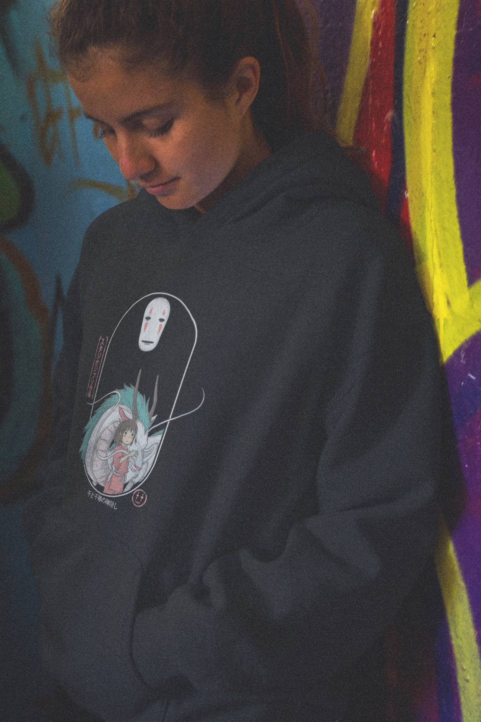 Person wearing a dark hoodie with a graphic design in front of colorful graffiti.
